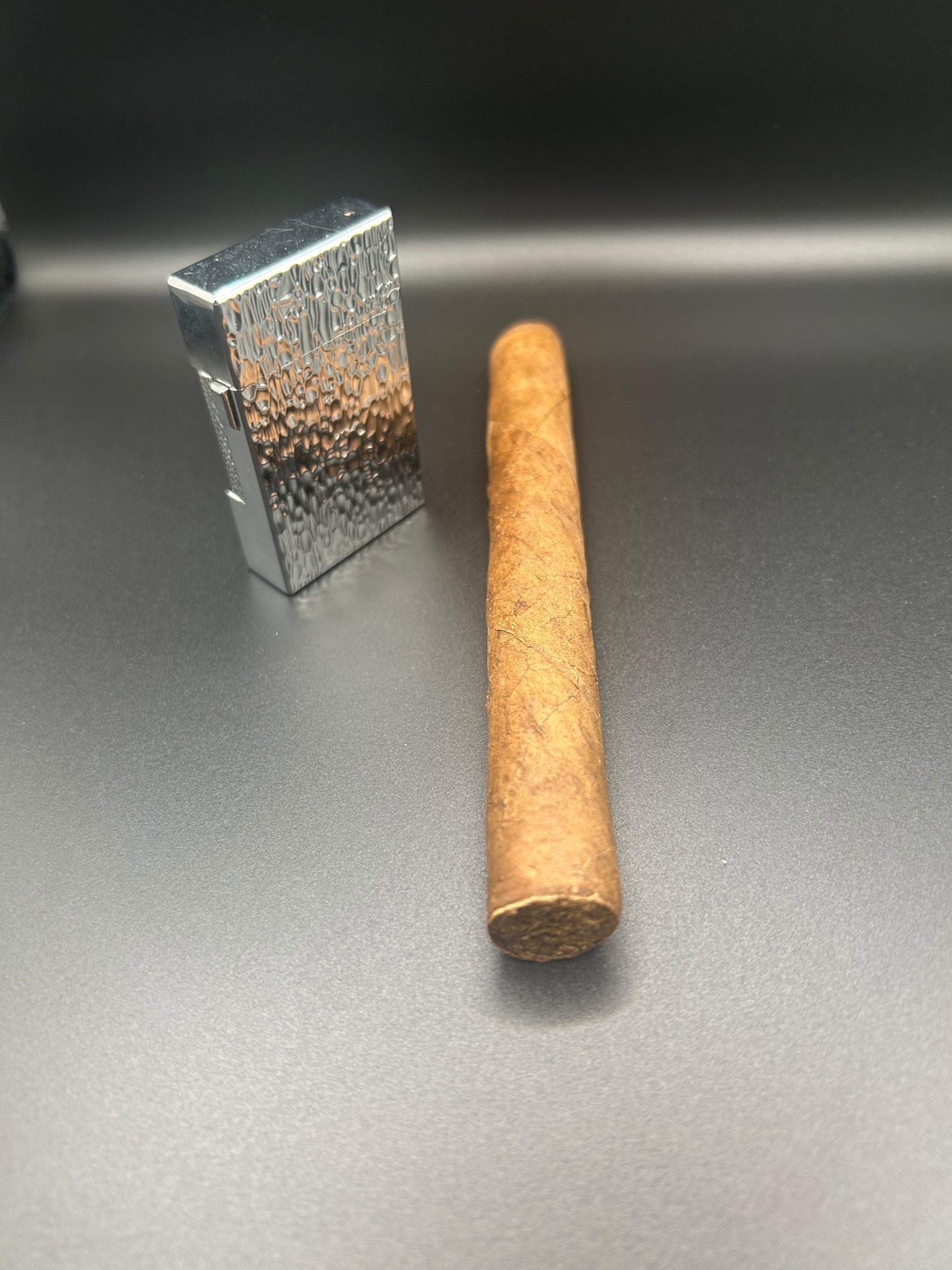 A silver, rectangular lighter and an unbranded cigar on a black background.